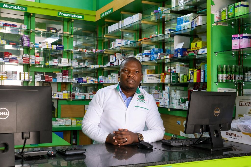 Given Pharmacy professional staff member at the counter, wearing a branded white jacket with hands clasped in a trustworthy, customer-ready pose, seated between dual Dell point-of-sale computers against a background of stocked green shelves featuring prescriptions, anti-malarials, and microbials.