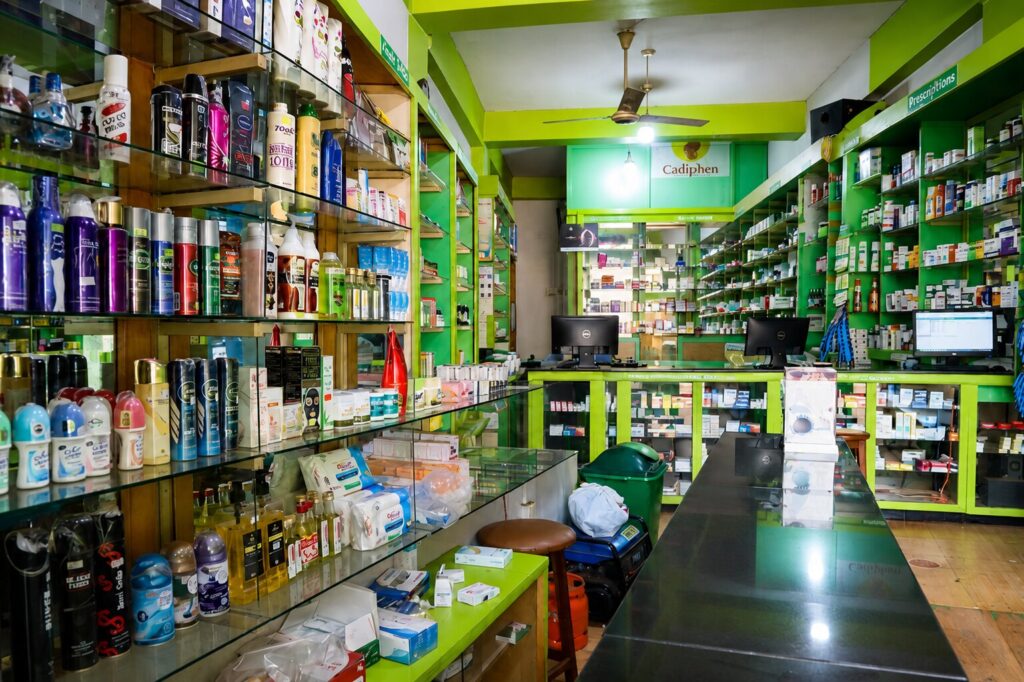 Interior view of Given Pharmacy's branch featuring brightly colored lime-green wood shelving stocked with a wide range of personal care toiletries and prescription pharmaceuticals, a glossy black service counter equipped with dual Dell computer monitors, and a green trash bin next to a round stool in a clean, organized retail space.