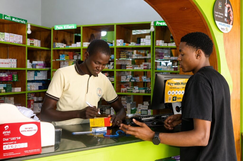 Given Pharmacy staff assisting a customer at the retail counter, writing on a medication box in front of well-organized shelves displaying anti-malarials, pain killers, and prescription items, with prominent Airtel Money and MoMo mobile payment terminals visible.
