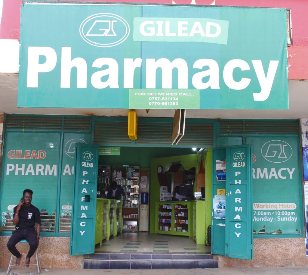 Exterior storefront of Given Pharmacy's Republic Street branch in Mbale, featuring a large green sign with delivery contact numbers, open folding doors with vertical branding, daily business hours clearly displayed from 7 am to 10 pm, and a man seated on a stool outside the entrance.