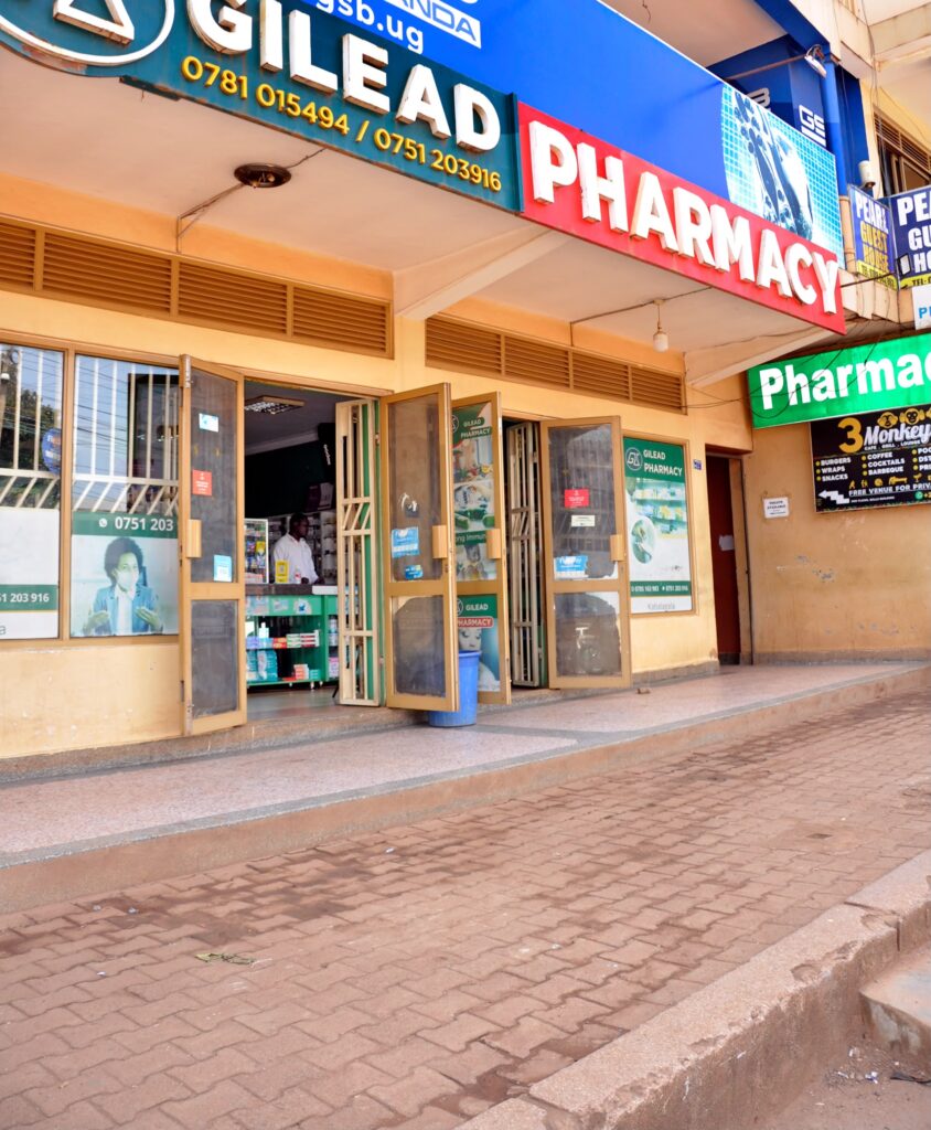 Exterior street view of Given Pharmacy's Kabalagala branch located on Muyenga Road in Kampala, featuring a prominent blue and red Gilead Pharmacy signboard with contact phone numbers, open double glass doors, and a paved brick sidewalk leading up to the entrance.