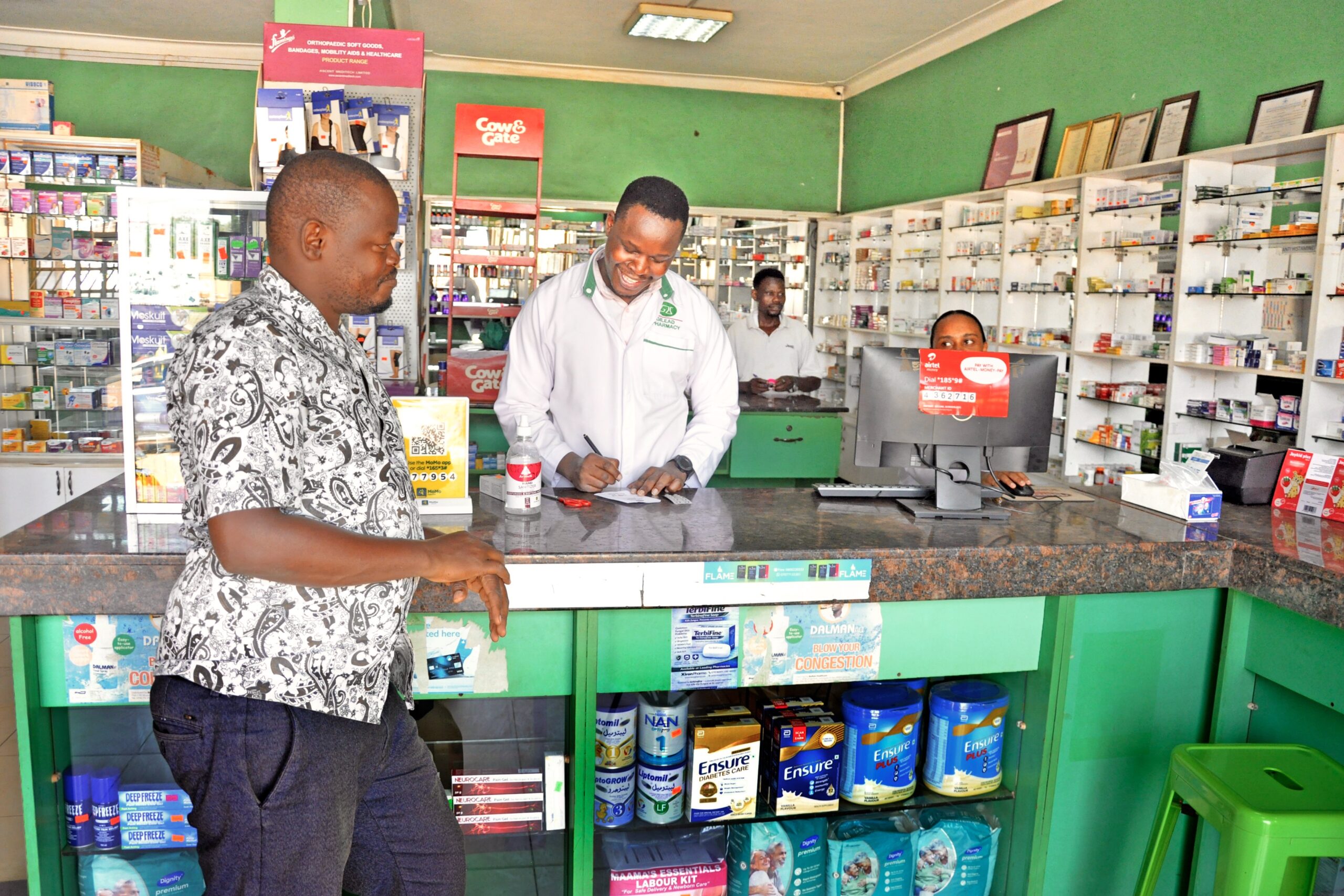 interior_kabalagala Given Pharmacy staff providing friendly customer service at the retail counter, featuring a smiling pharmacist assisting a male customer, surrounded by fully stocked green shelves displaying baby formula (NAN, Cow & Gate), nutritional supplements (Ensure), and pain relief patches.