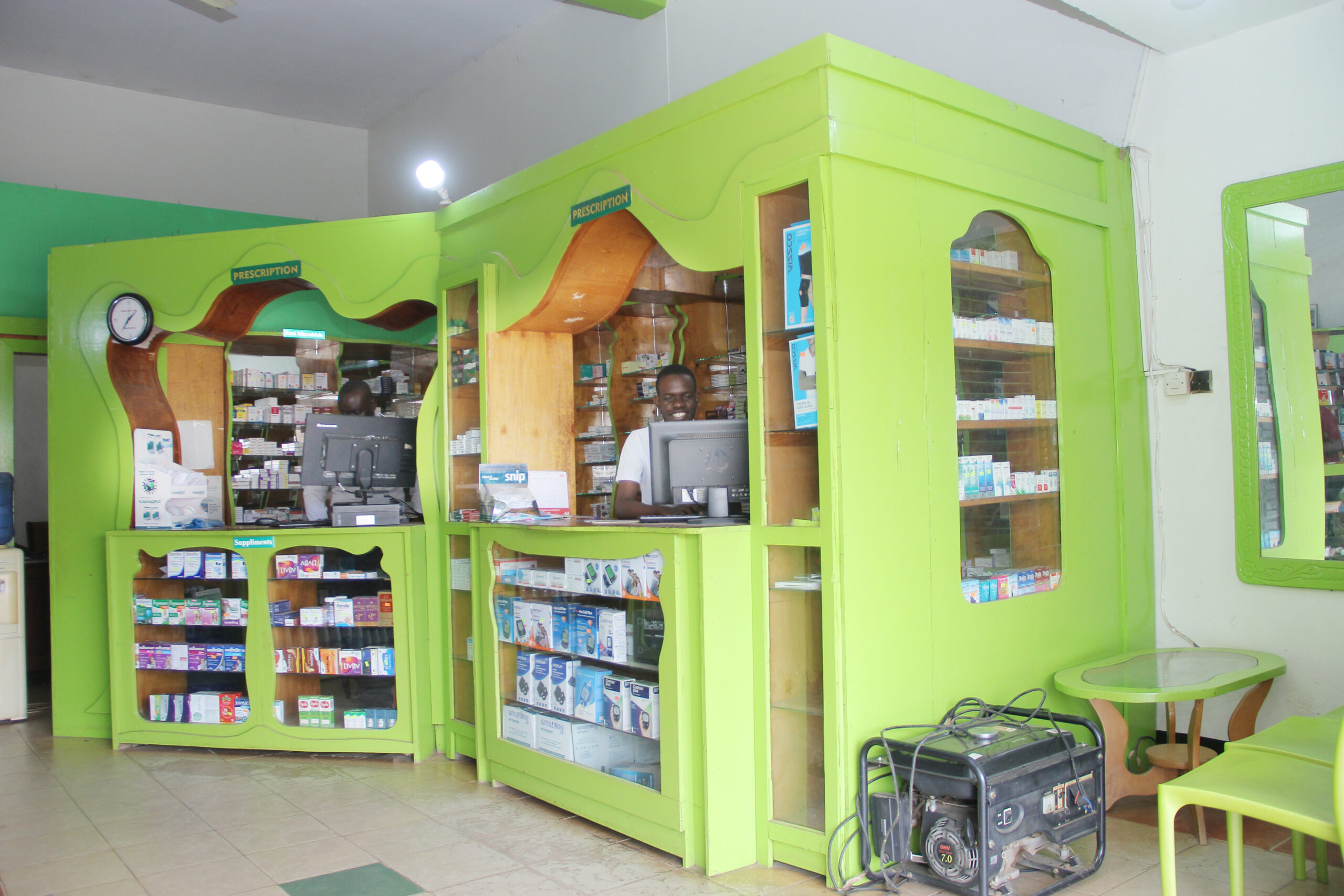 Interior view of Given Pharmacy's Republic Street Mbale branch showcasing the signature lime-green wavy wooden prescription counters and glass display cabinets, with a smiling staff member working behind the computer monitor, alongside a portable generator, waiting chairs, and a wall mirror reflecting the bright, clean space.