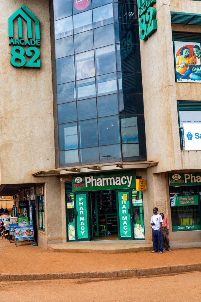 Exterior storefront of Given Pharmacy's Mainstreet branch located at the Arcade 82 building, featuring the brand's distinctive green signage, adjacent Given Optics shop offering free eye check-ups, and pedestrians walking on the sidewalk outside the entrance.