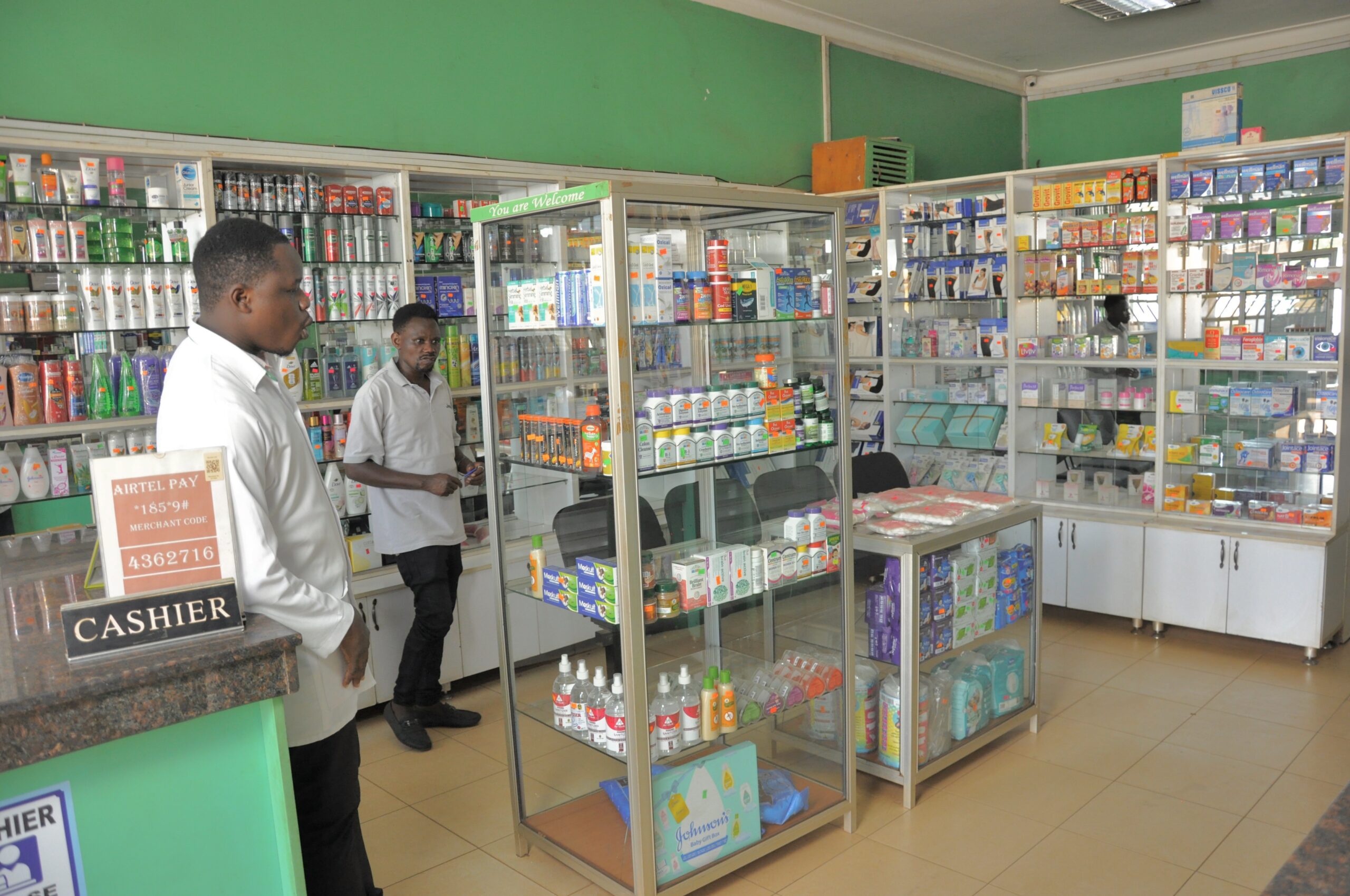 well_stocked_kabalagala Interior view of Given Pharmacy's Kabalagala branch showing a staff member standing near the black cashier counter alongside prominent Airtel Pay mobile money merchant signage, with a central glass display cabinet, a 'You are Welcome' greeting sign, and white shelving units fully stocked with pharmaceuticals, personal care items, and baby care products.