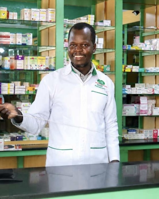 Given Pharmacy chronic care specialist and pharmacist smiling warmly behind the sleek black service counter, wearing a branded white lab coat with the Given Pharmacy logo, set against the signature green wooden shelving stocked with a wide range of medications.